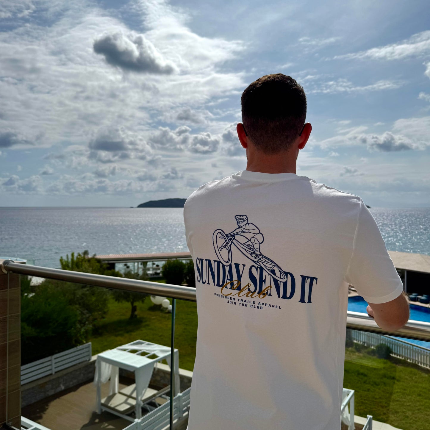 Person wearing a white t-shirt with 'SUNDAY SEND IT' text, standing on a balcony overlooking water and sky.