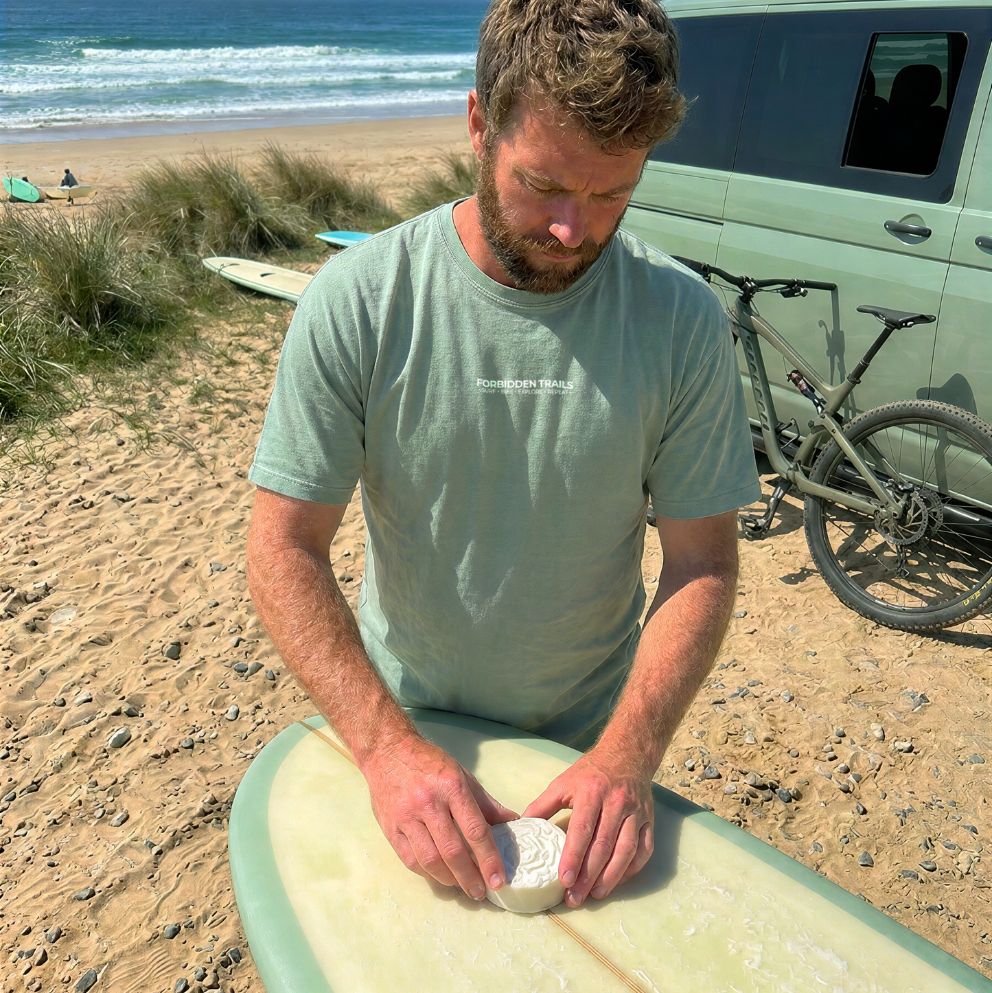 A man on a sandy beach preparing a surfboard, wearing a light sage green t-shirt and working with a light green surfboard in front of a van and ocean.