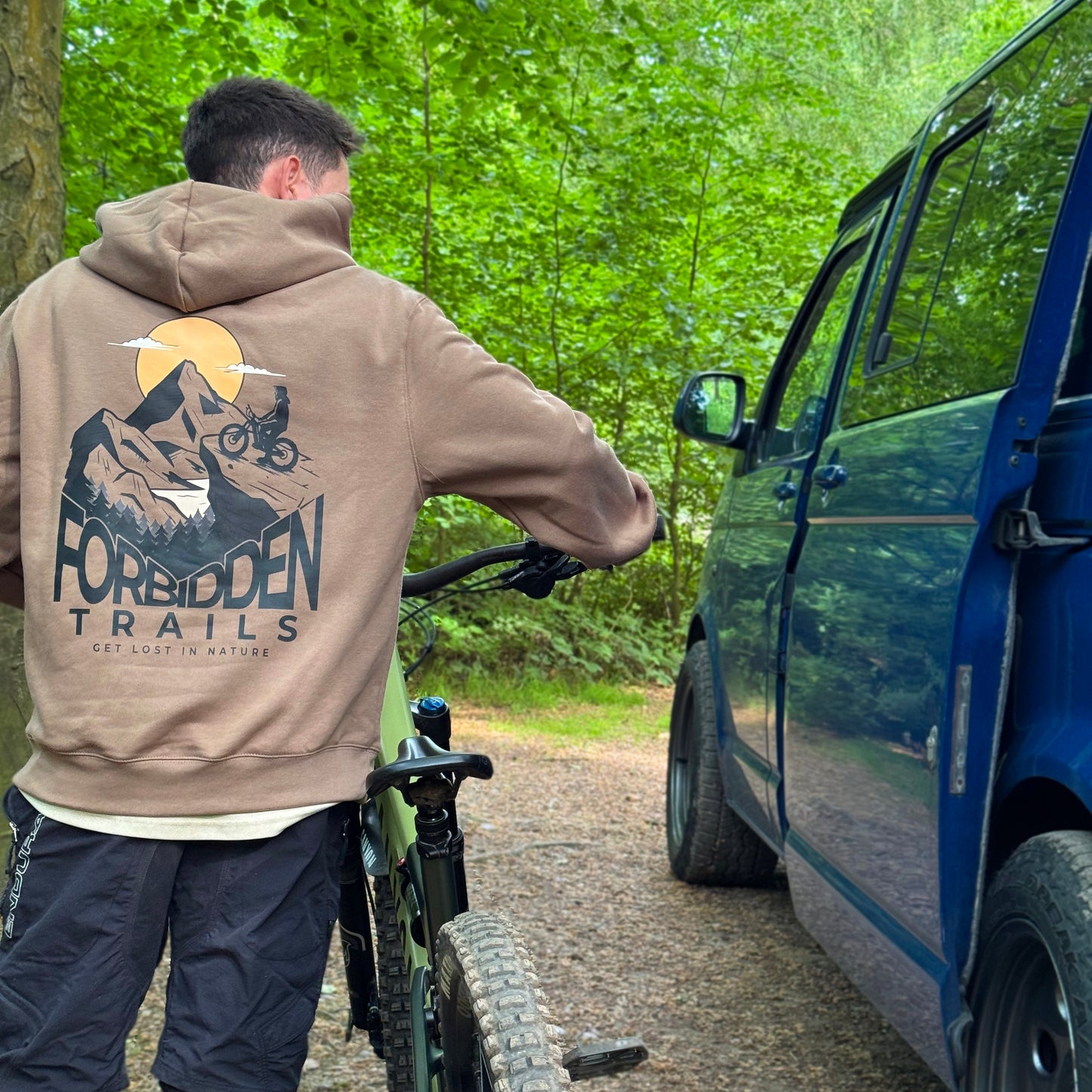 Person with 'Forbidden Trails' hoodie standing next to a bike and vehicle in a forest.