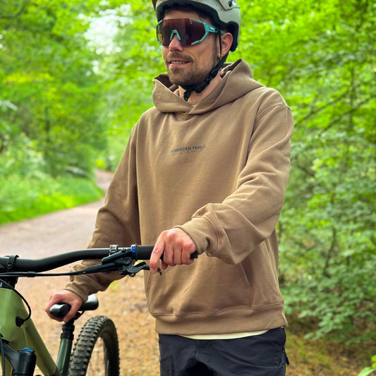 Man with a bicycle in a forested area wearing a brown hoodie and helmet.