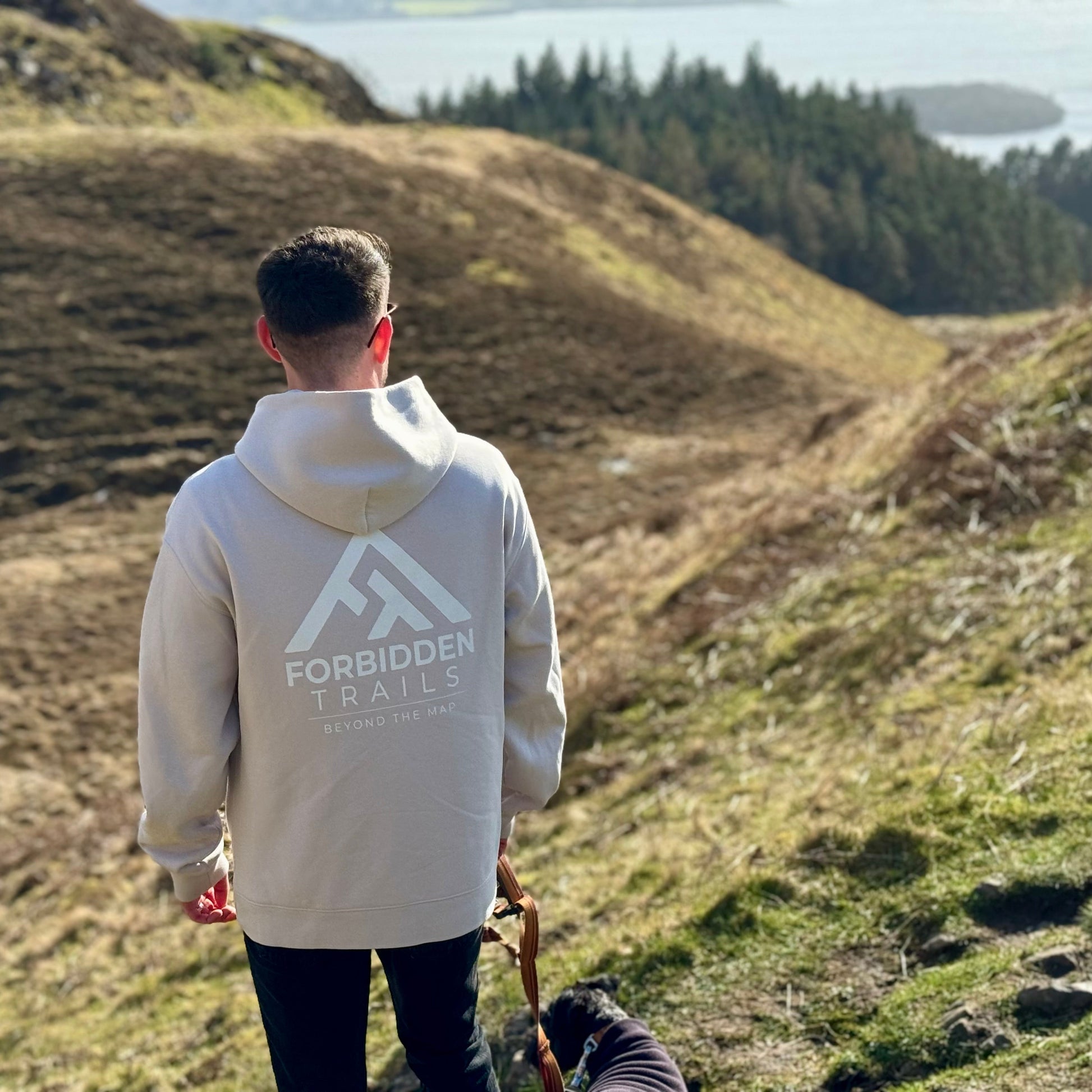 Person wearing a hoodie with 'Forbidden Trails' logo walking on a grassy hillside.