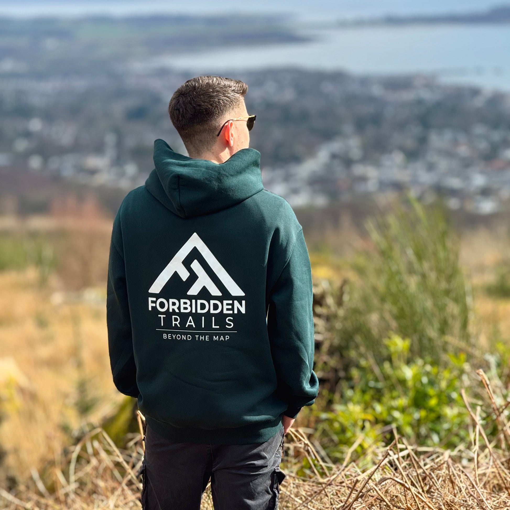 Person wearing a hoodie with 'Forbidden Trails' logo overlooking a scenic landscape.