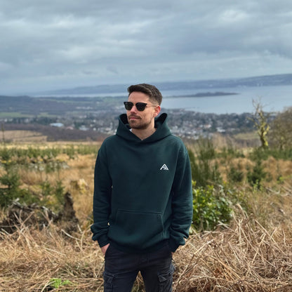 Man wearing a dark green hoodie with a logo, standing in a field with a scenic background.