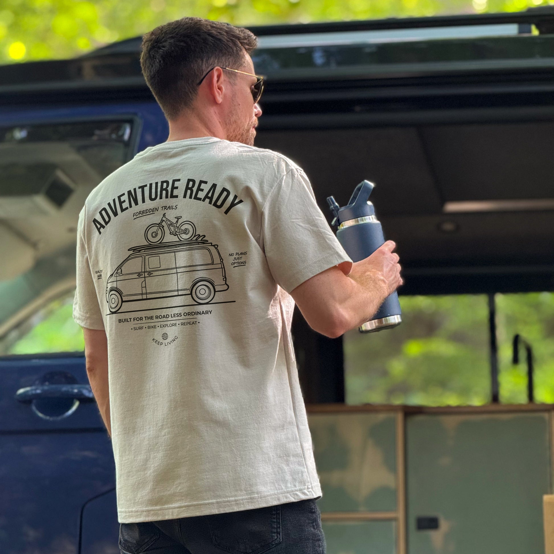 Man wearing an 'Adventure Ready' t-shirt holding a water bottle outdoors.
