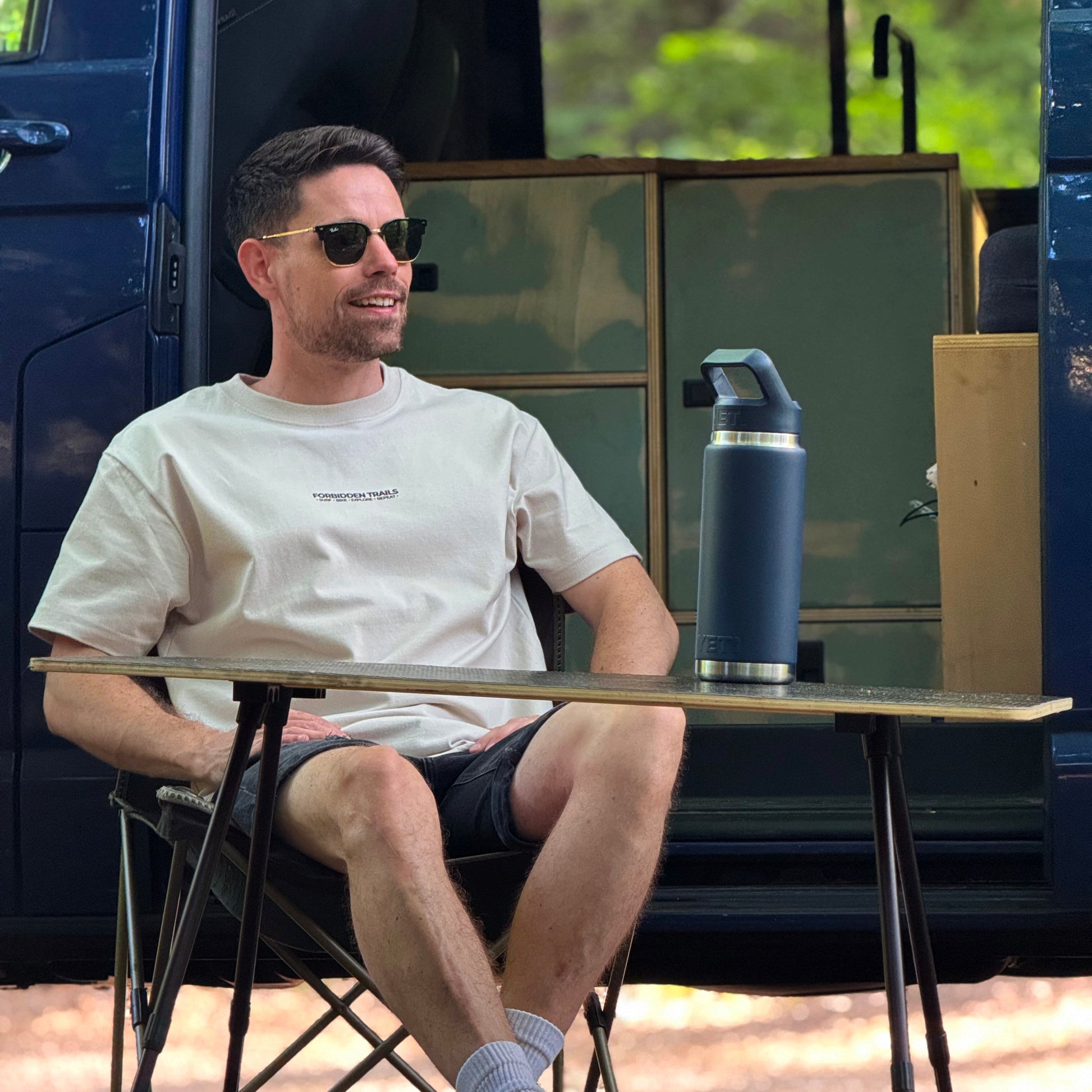 Man sitting at a table outdoors with a blue thermos and a van in the background