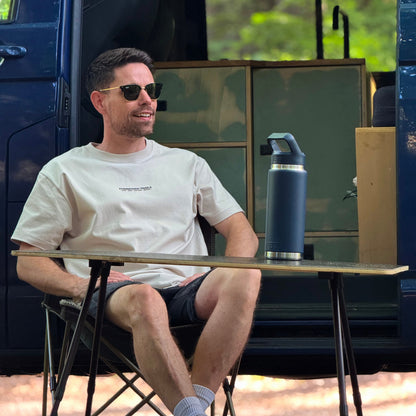 Man sitting at a table outdoors with a blue thermos and a van in the background