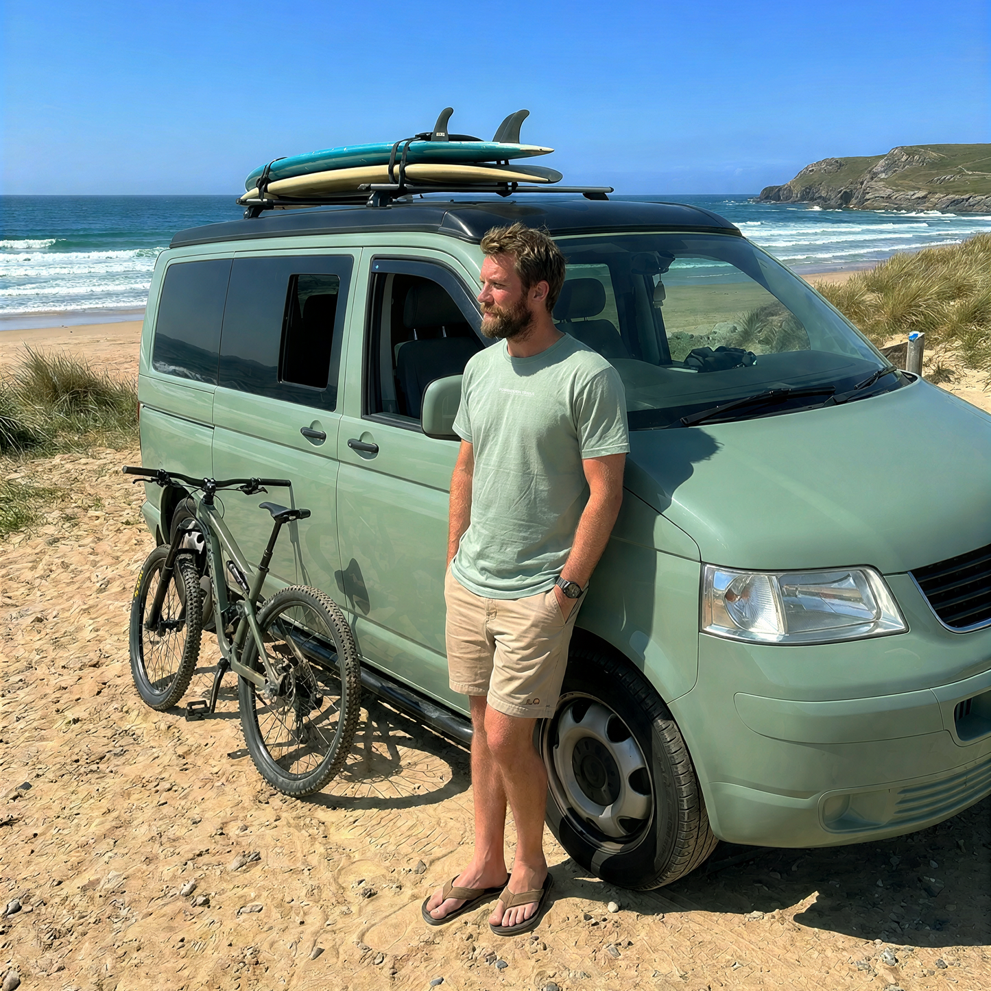 A man stands on a sandy beach next to a sage green Volkswagen Transporter van with surfboards on the roof rack and a bicycle parked beside it, with the ocean and blue sky in the background.