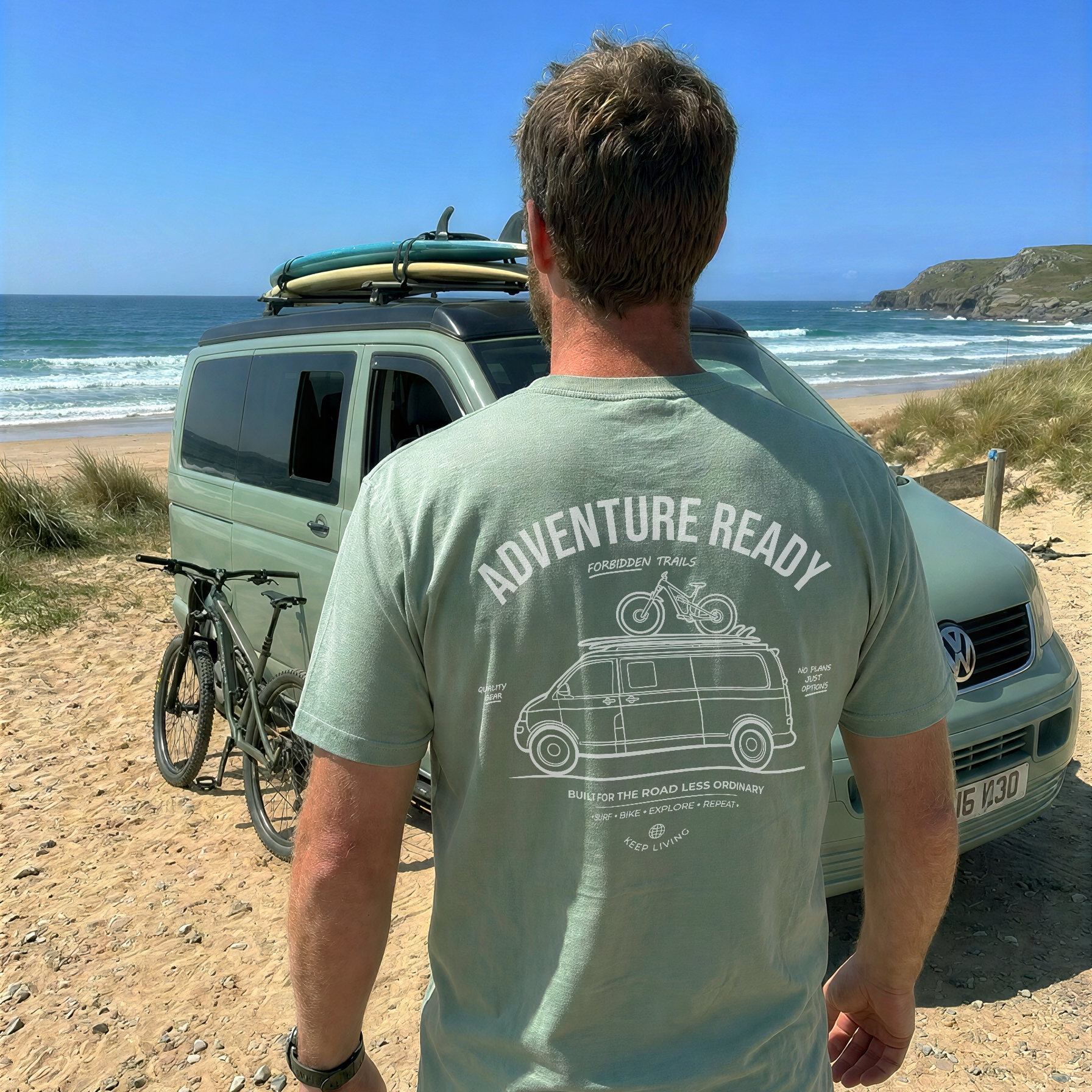 A man standing on a sandy beach facing a van with a surfboard on top, wearing a sage green t-shirt with a white graphic print that reads 'ADVENTURE READY'.