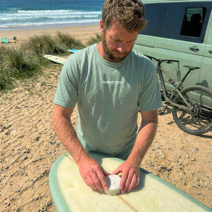 A man on a sandy beach preparing a surfboard, wearing a light sage green t-shirt and working with a light green surfboard in front of a van and ocean.