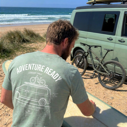 A man on a sandy beach, seen from behind, wearing a sage green t-shirt with a white graphic print of a camper van and the words 'ADVENTURE READY'. He is standing next to a surfboard, with a green camper van, a bicycle, and the ocean in the background.