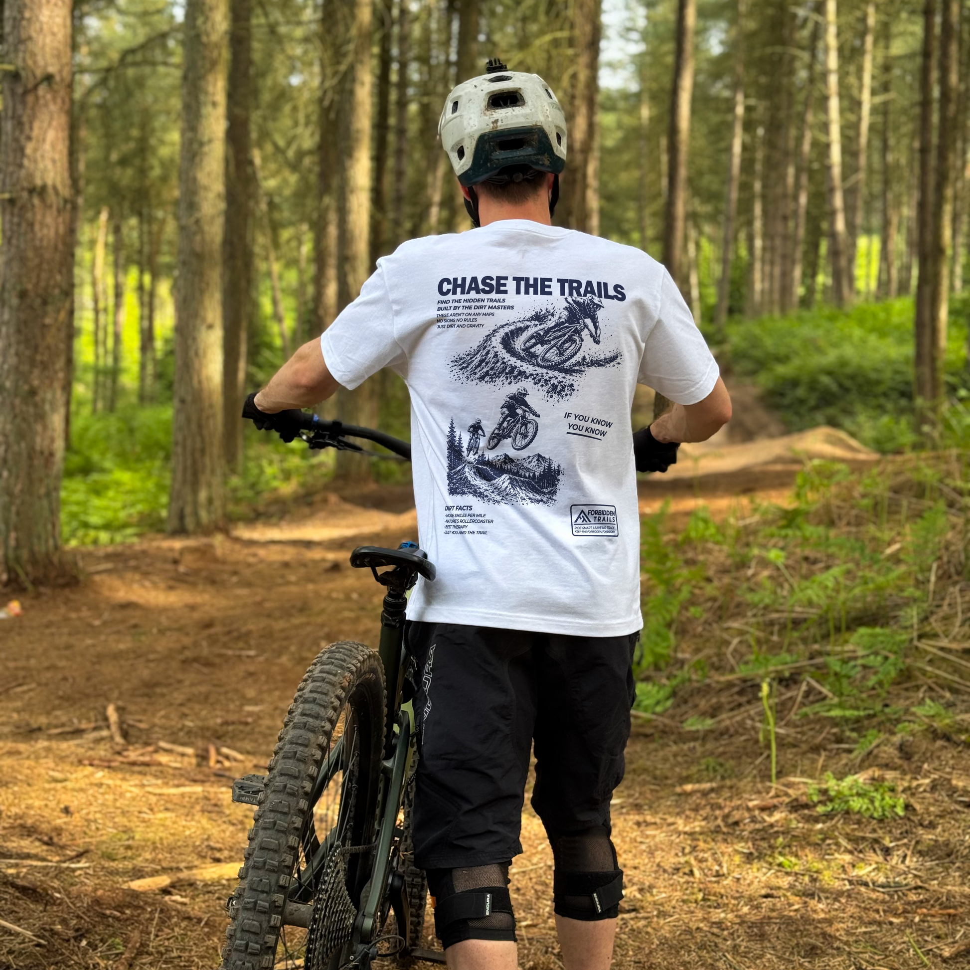 Person with a mountain bike in a forest wearing a white t-shirt with graphics and text.