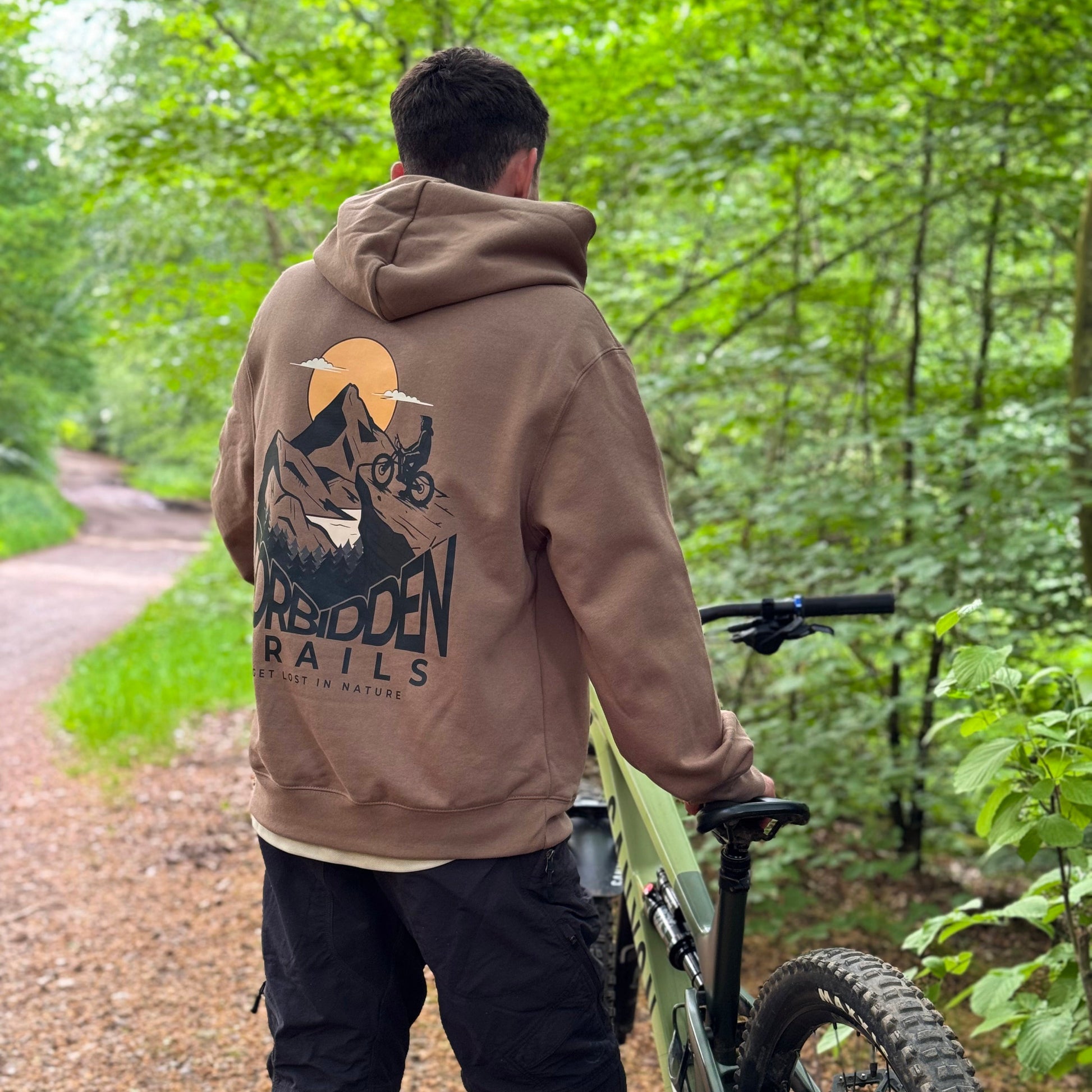 Person wearing a hoodie with a graphic design, standing in a forest with a bike.