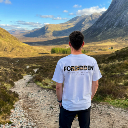 Person standing on a mountain path wearing a white t-shirt with 'FORBIDDEN' text, looking at scenic mountains.
