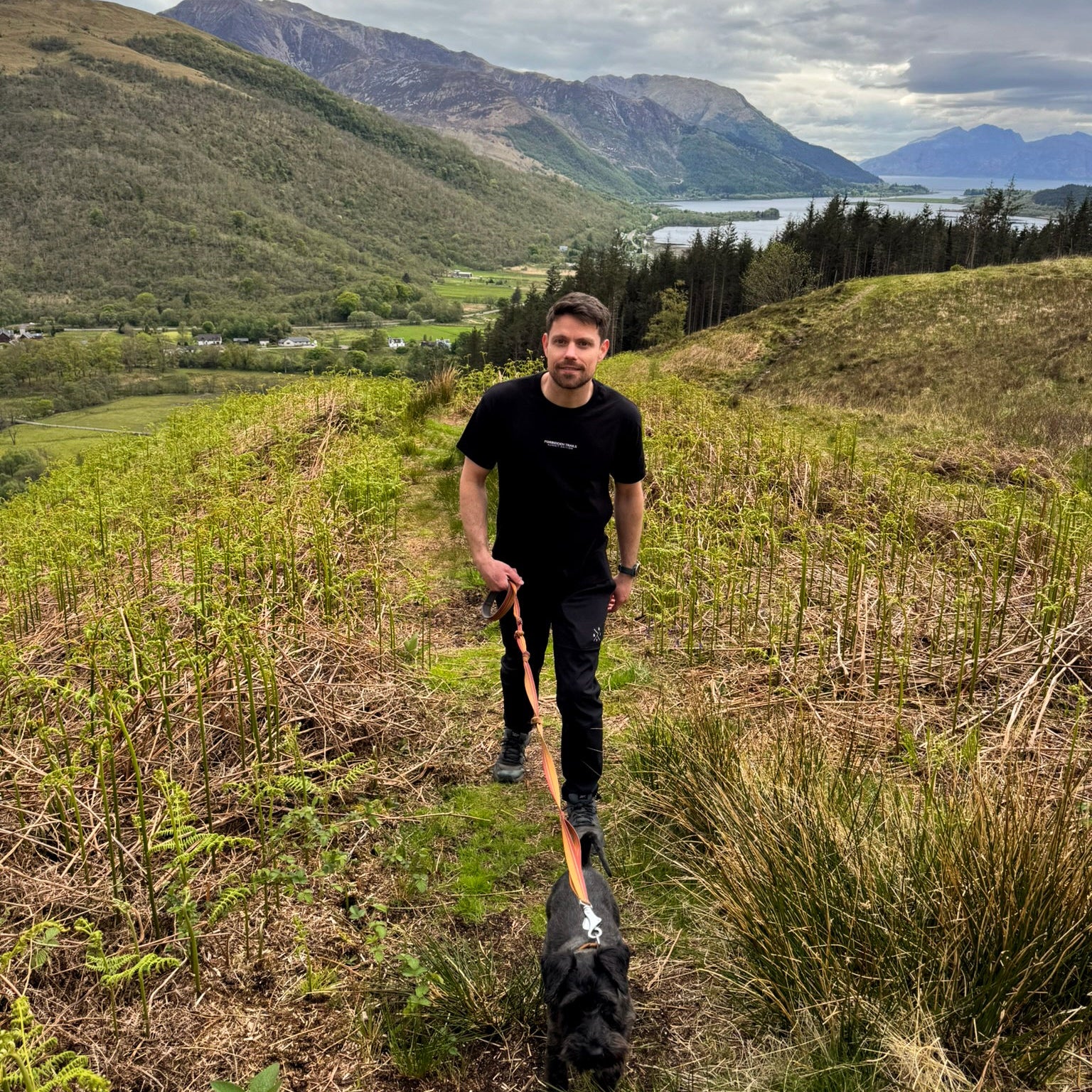 man walking up hill with dog mountain and lakes in the background