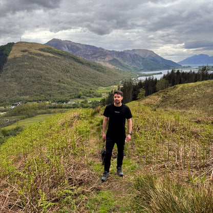 Man standing on a grassy hill with mountains and a lake in the background